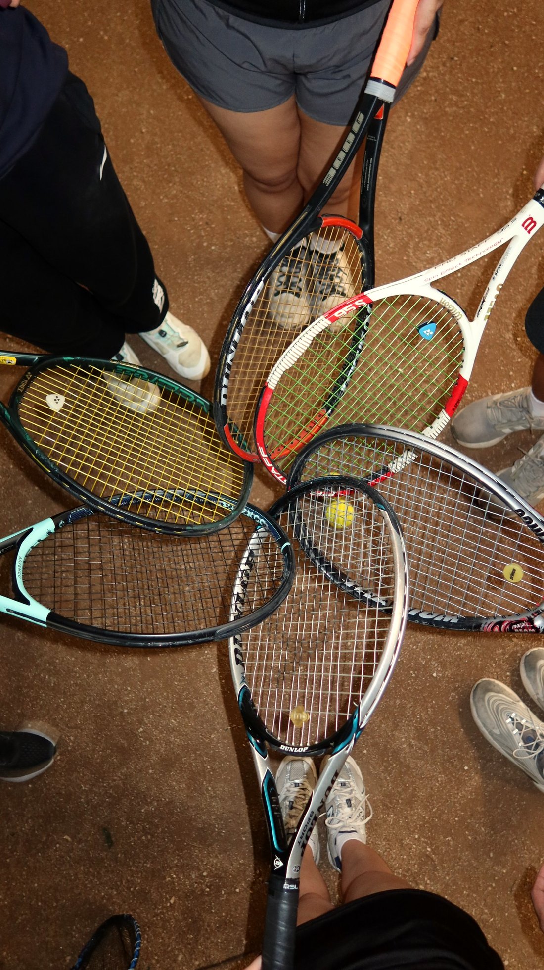 Seen from above: six friends on a clay court touch their rackets together in a circle, frames from Yonex, Wilson, Dunlop, Prince and Artengo