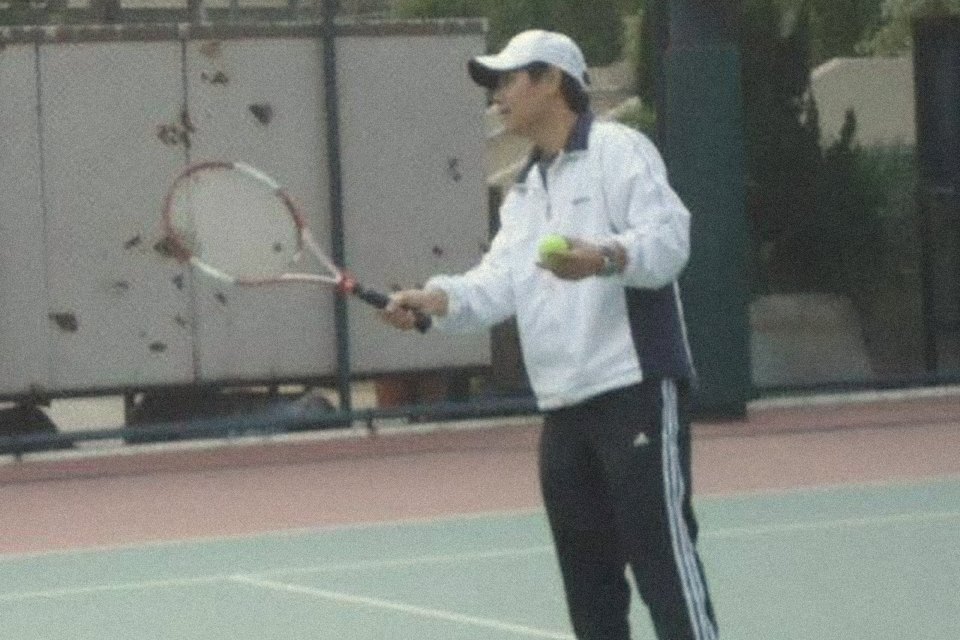 The coach in a white track jacket and cap, feeding a ball on a Hong Kong hard court
