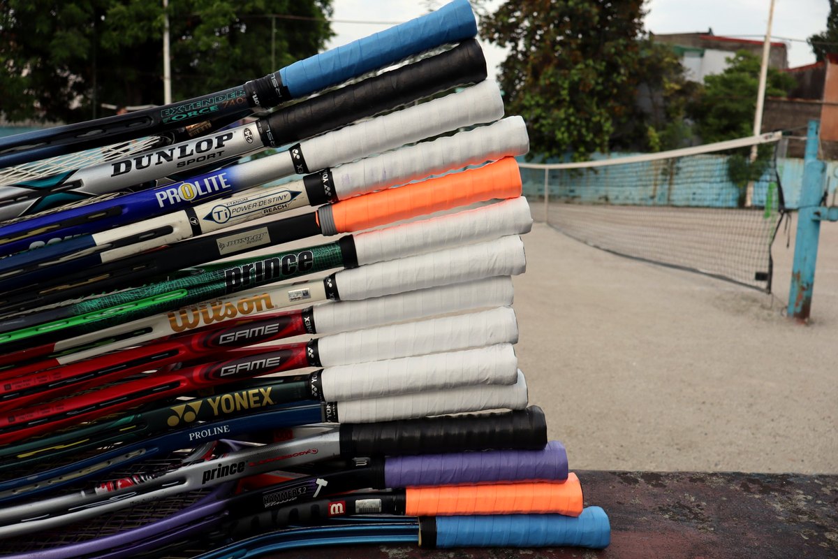 A stack of rental rackets by their grips, Dunlop, Prince, Wilson and Yonex frames, beside an open court