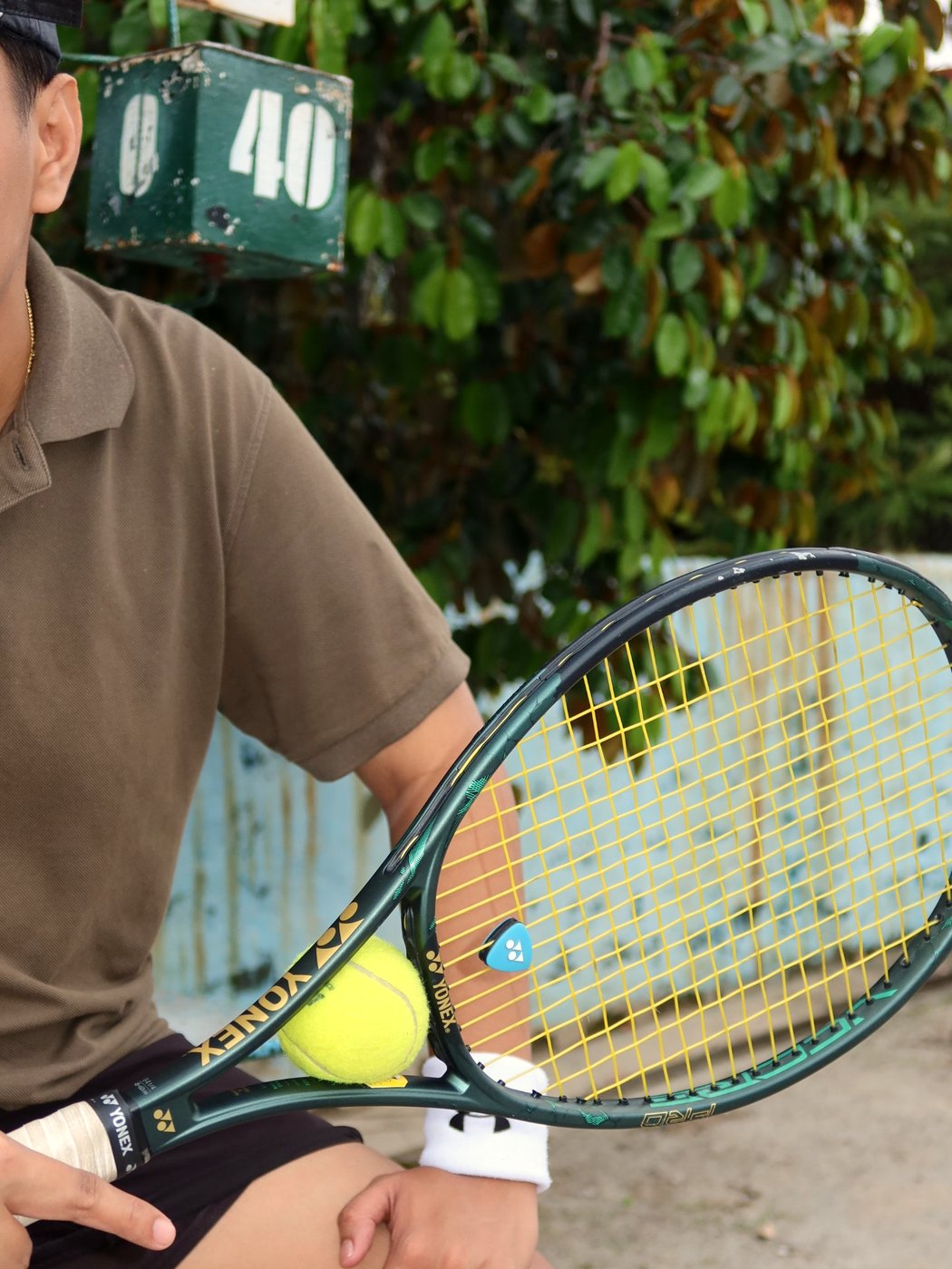 A player in an olive polo sits courtside by the old green scoreboard, a Yonex VCORE PRO resting on his knee with a ball tucked in its throat