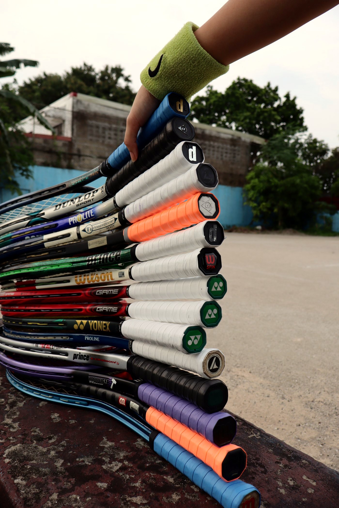 One arm in a lime sweatband holding fifteen rental rackets by their grips, the frames fanned out on a weathered ledge beside the court