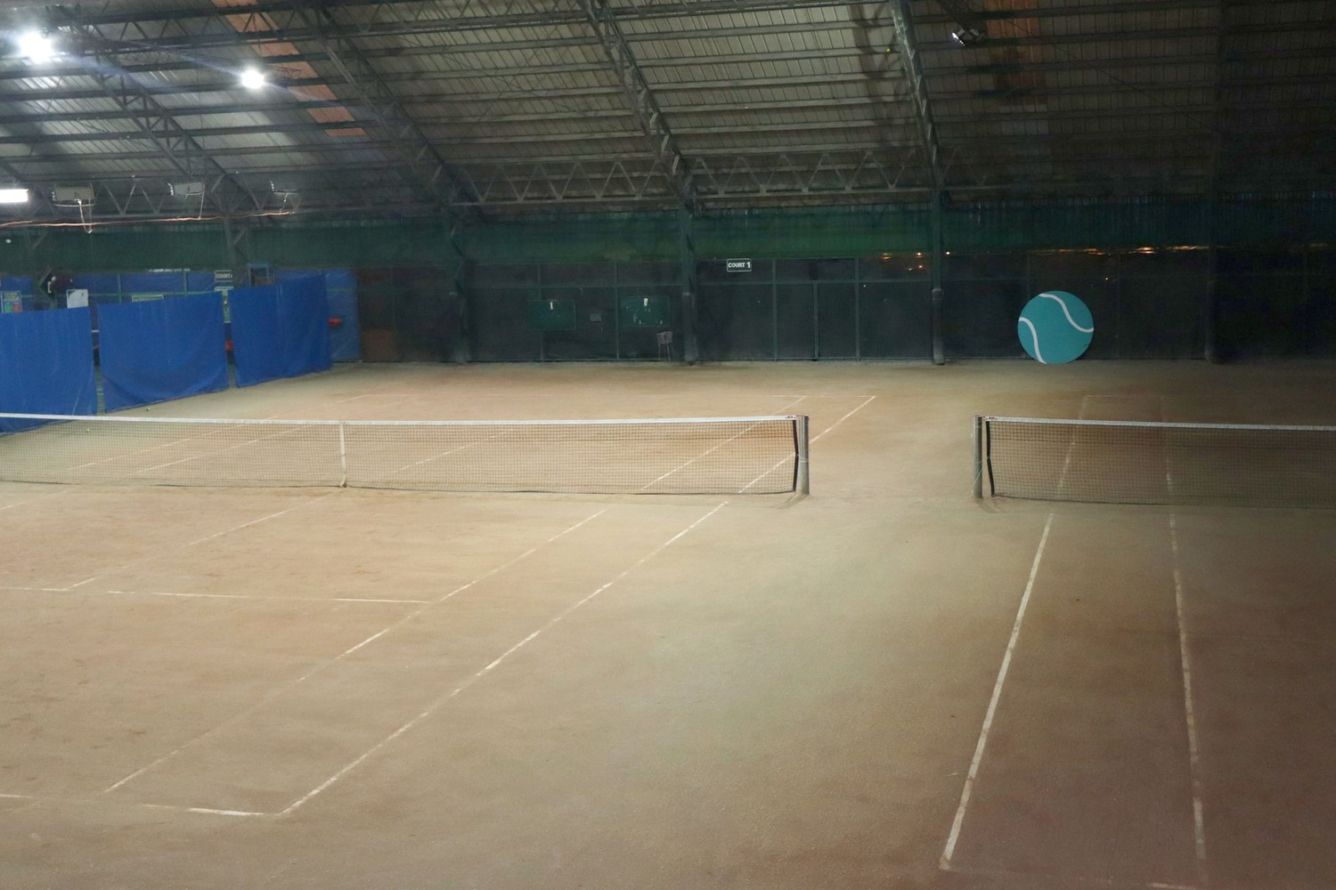 The indoor clay courts under the roof trusses, two nets, a COURT 1 sign and a large teal tennis ball painted on the back wall