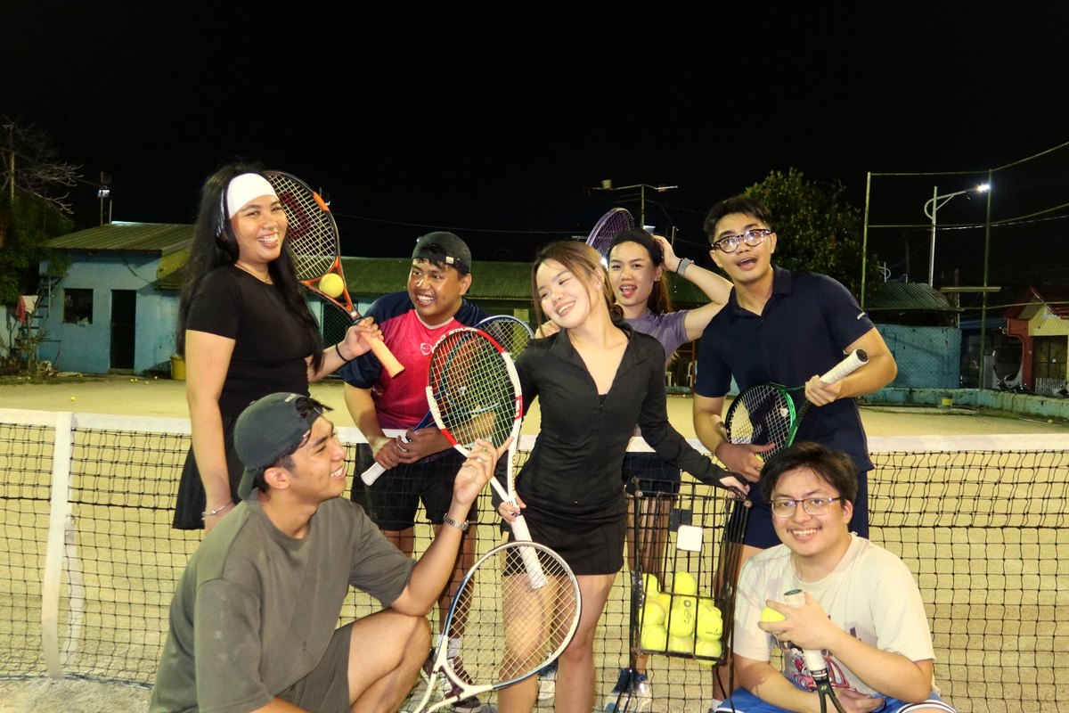 Seven friends laughing at the net on a lit court at night, rackets up, a basket of balls between them