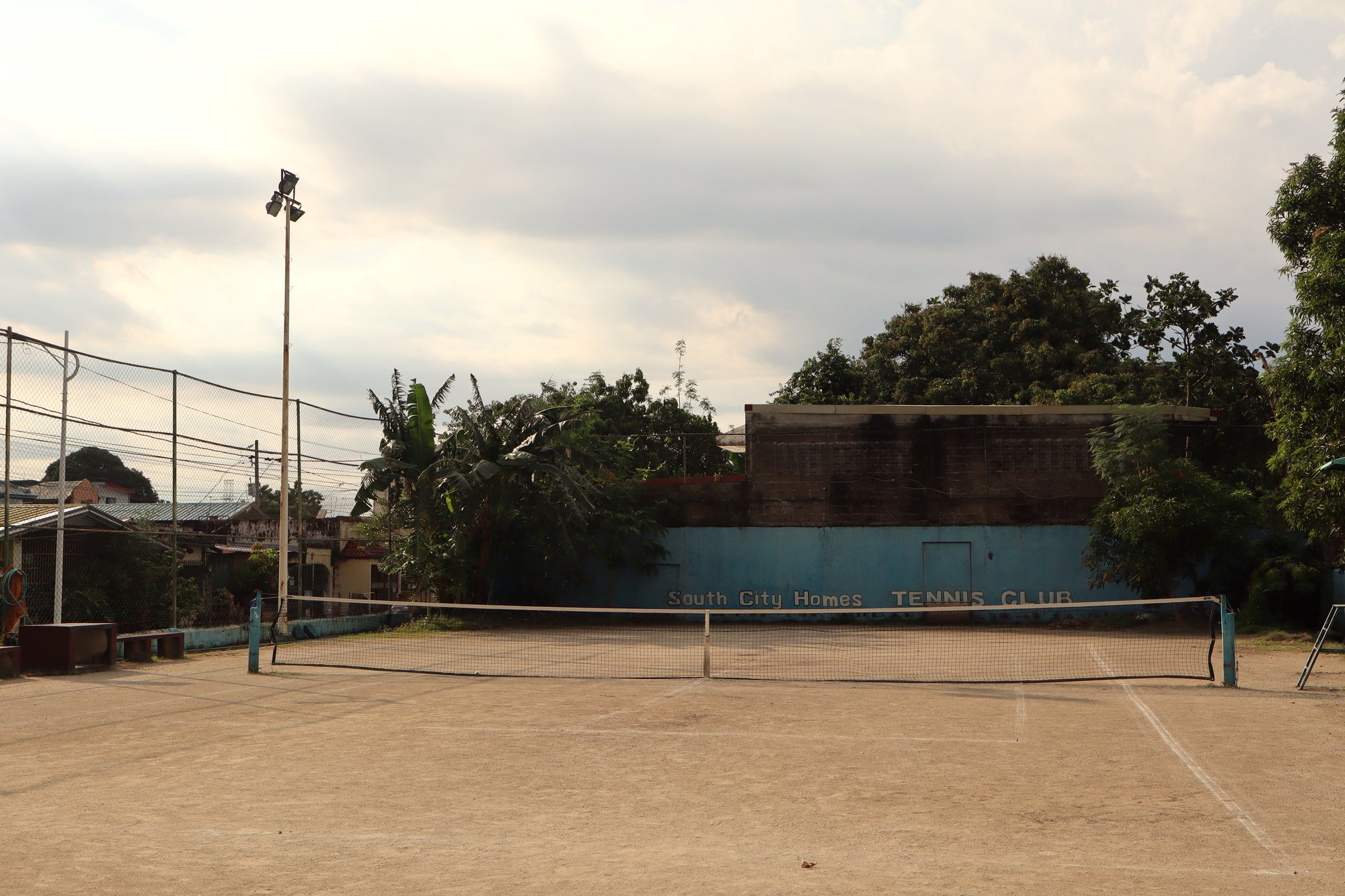The outdoor court at South City Homes in daylight: the net, a floodlight pole on the left, and the blue wall painted South City Homes Tennis Club under a cloudy sky